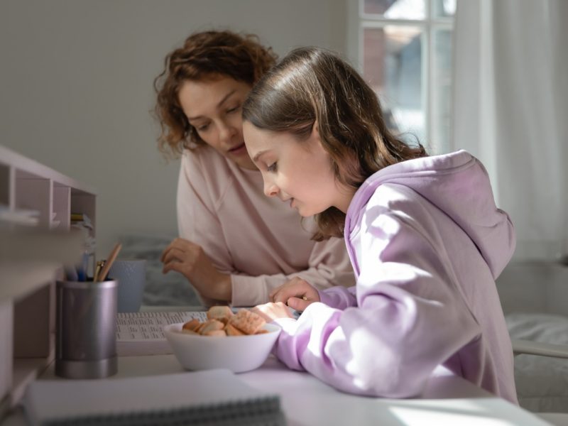 Focused mom helping teenage daughter doing homework studying from home. Young adult parent mum or tutor, coach teaching school child, explaining distance learning education reading book together.